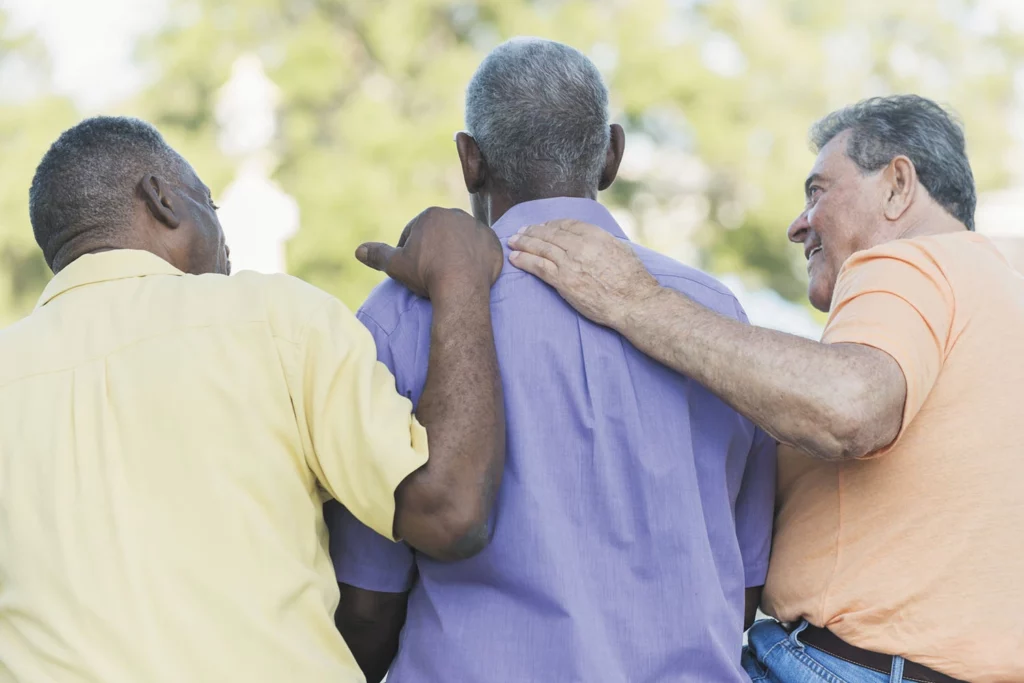 Three men are seen from behind, walking together outdoors. One man in a yellow shirt, one in a purple shirt, and another in an orange shirt. They are engaging warmly, with hands on each other's shoulders, suggesting friendship and support. Trees and greenery are visible in the background.